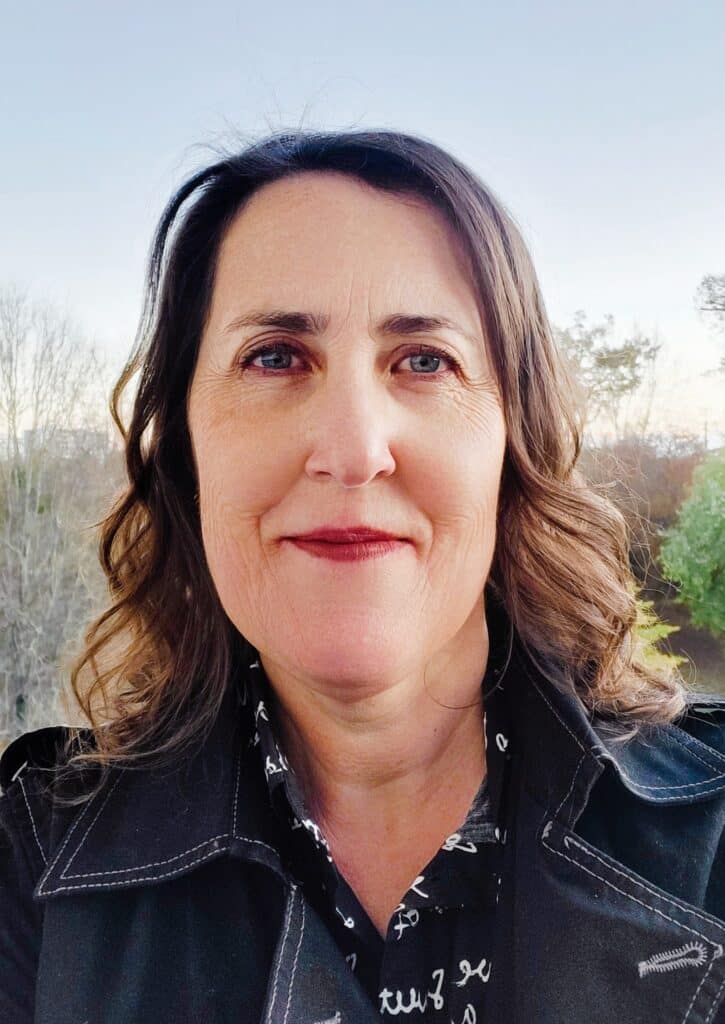 Femme mature et confiante : portrait extérieur élégant. Portrait extérieur d'une femme mature, cheveux bruns ondulés. Elle sourit, yeux bleus, manteau noir à coutures blanches, fond nature flou.